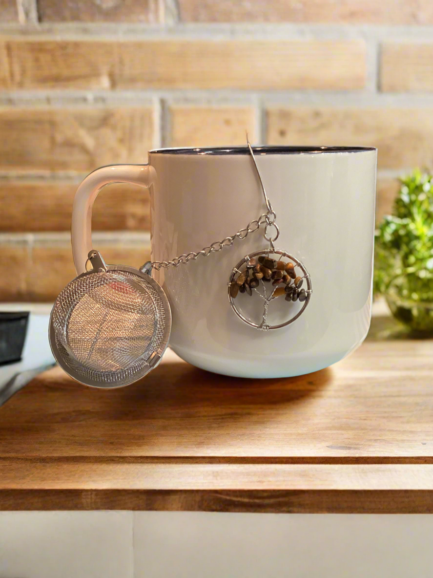 A white ceramic tea cup with a silver mesh tea strainer attached to it, featuring a tree of life design with a tiger's eye stone, placed on a wooden surface.