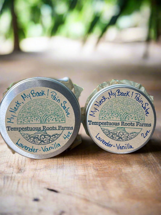 A small open aluminum tin containing green salve, with a lid featuring text that includes 'My Neck, My Back!!' and mentions of lavender and vanilla. The tin is placed on a wooden surface with a sprig of lavender and vanilla pods scattered around.