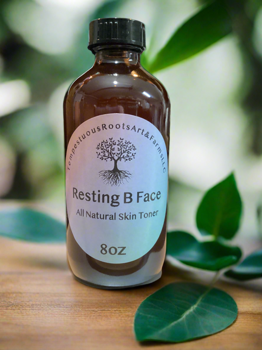 Bottle of 'Resting B Face' all natural skin toner with a label on a wooden surface and green leaves in the background.