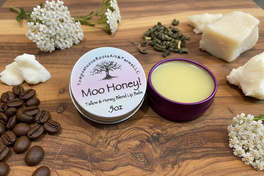 Moo Honey! Tallow & Honey Lip Balm
 by Tempestuous Roots Art & Farms LLC on a wooden surface with coffee beans, butters, and herbs about