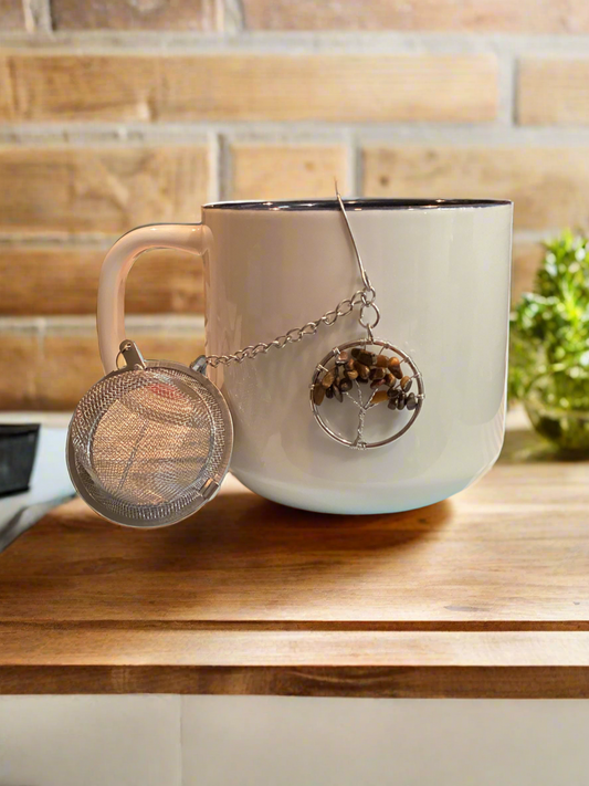 A white ceramic tea cup with a silver mesh tea strainer attached to it, featuring a tree of life design with a tiger's eye stone, placed on a wooden surface.